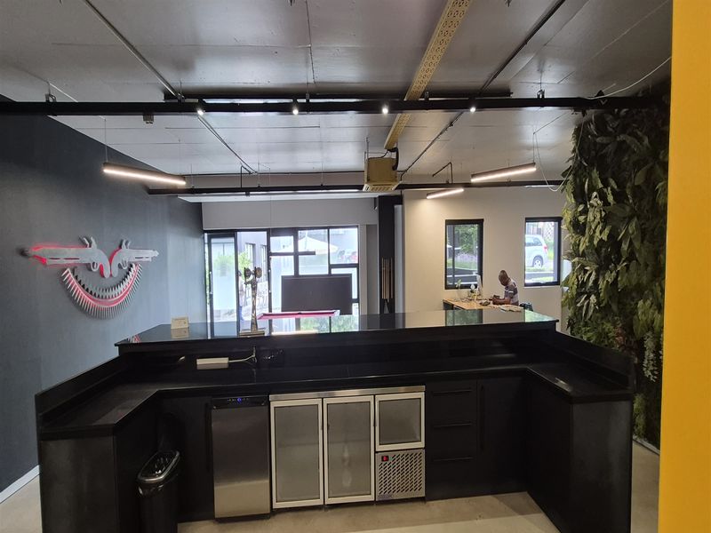 Modern office bar area featuring a sleek black counter, integrated under-counter refrigeration, track lighting, and a decorative vertical garden wall.