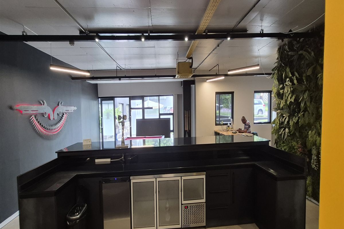 Modern office bar area featuring a sleek black counter, integrated under-counter refrigeration, track lighting, and a decorative vertical garden wall.