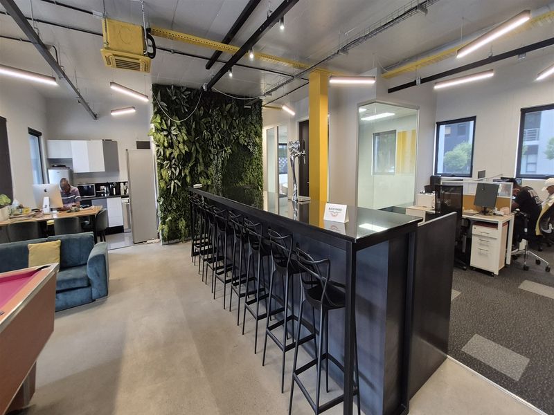 Modern open-plan office space featuring a central bar area, vertical garden wall, exposed industrial ceiling, and integrated kitchenette facilities.