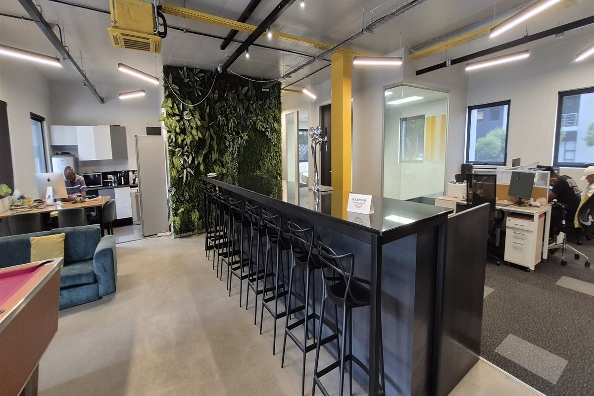 Modern open-plan office space featuring a central bar area, vertical garden wall, exposed industrial ceiling, and integrated kitchenette facilities.