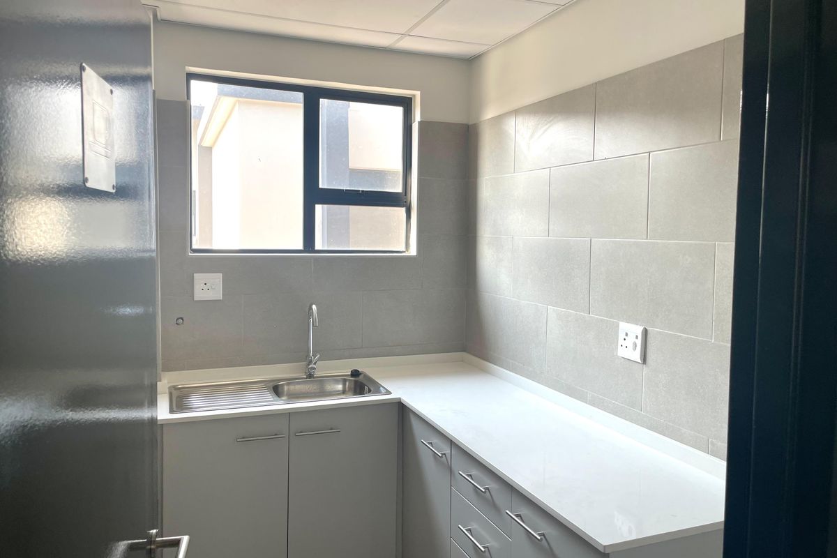 A clean, modern kitchenette featuring grey cabinetry, a stainless steel sink, white countertops, and neutral tiled backsplash in an office environment.