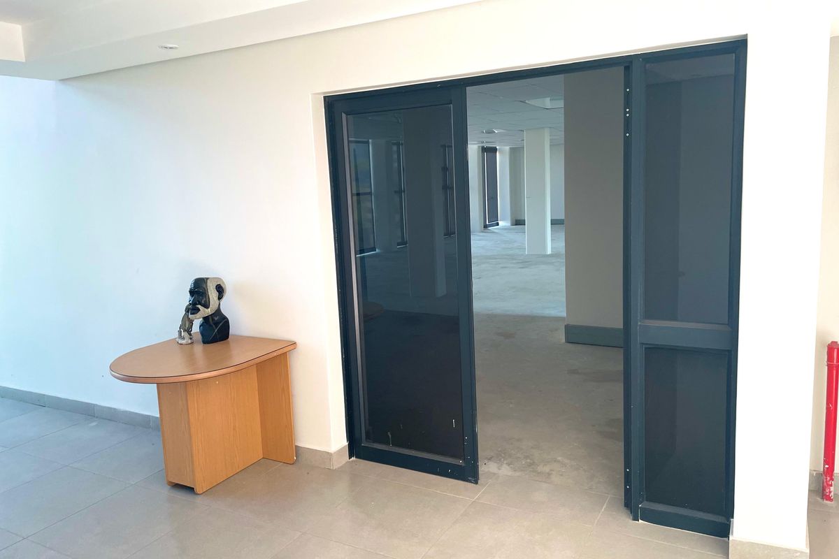 A bright, open-plan office space featuring neutral tiled flooring, recessed lighting, and a modern aluminium-framed glass entrance door.