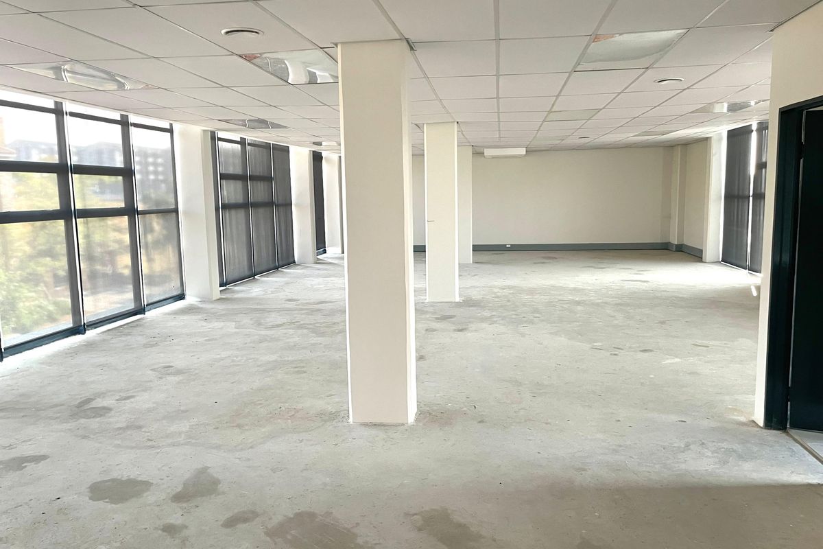 An open-plan office space featuring large windows, structural columns, and a suspended ceiling, currently in a vacant, shell-like condition.