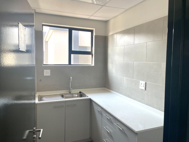 A clean, modern kitchenette featuring grey cabinetry, a stainless steel sink, white countertops, and neutral tiled backsplash in an office environment.