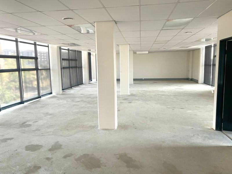 An open-plan office space featuring large windows, structural columns, and a suspended ceiling, currently in a vacant, shell-like condition.
