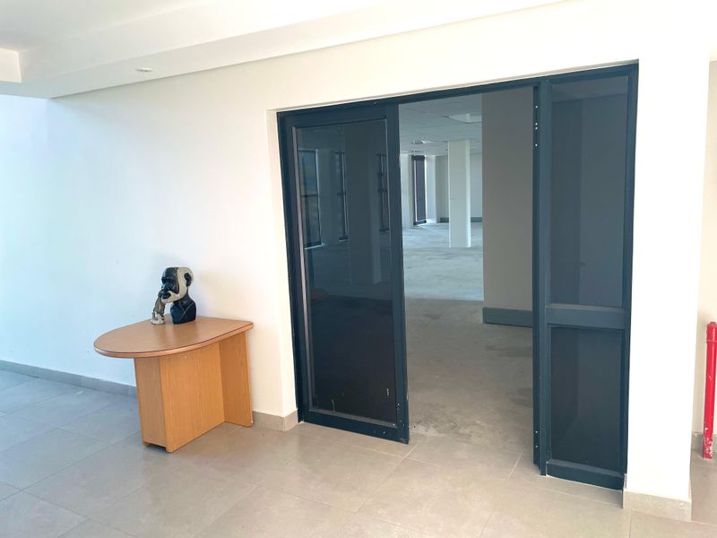 A bright, open-plan office space featuring neutral tiled flooring, recessed lighting, and a modern aluminium-framed glass entrance door.