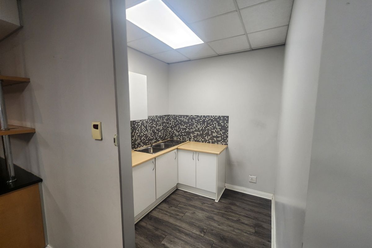 A compact office kitchenette featuring white cabinetry, a double sink, mosaic tile backsplash, and wood-look flooring under a drop ceiling.