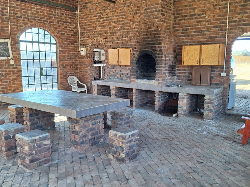 Spacious covered outdoor patio featuring rustic brick construction, a built-in braai, integrated brick dining table, and large arched windows overlooking the farm landscape.
