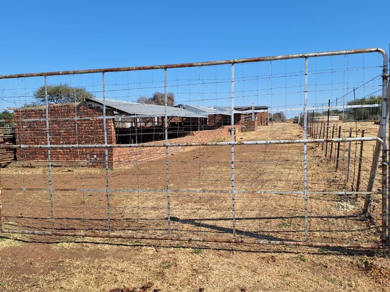 A view of a farm property featuring a metal gate, wire fencing, and a brick livestock shelter with a corrugated metal roof.
