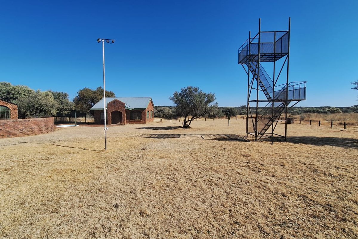 Expansive farm grounds featuring a brick building, a metal observation tower, and a floodlight pole set within a dry, open landscape.