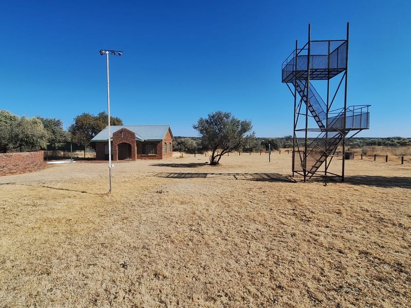 Expansive farm grounds featuring a brick building, a metal observation tower, and a floodlight pole set within a dry, open landscape.