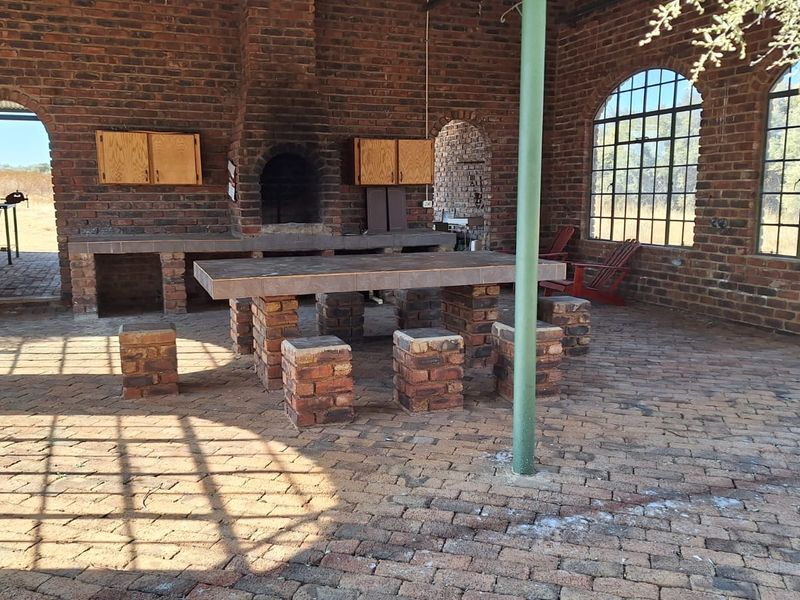 Spacious covered outdoor patio featuring rustic exposed brick walls, a built-in fireplace, brick-paved flooring, and large arched windows overlooking the farm landscape.