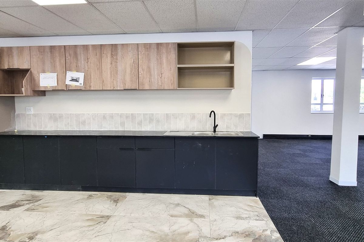 A modern office kitchenette featuring dark base cabinetry, wood-look overhead cupboards, a stainless steel sink, and tiled flooring, set within an open-plan office space.