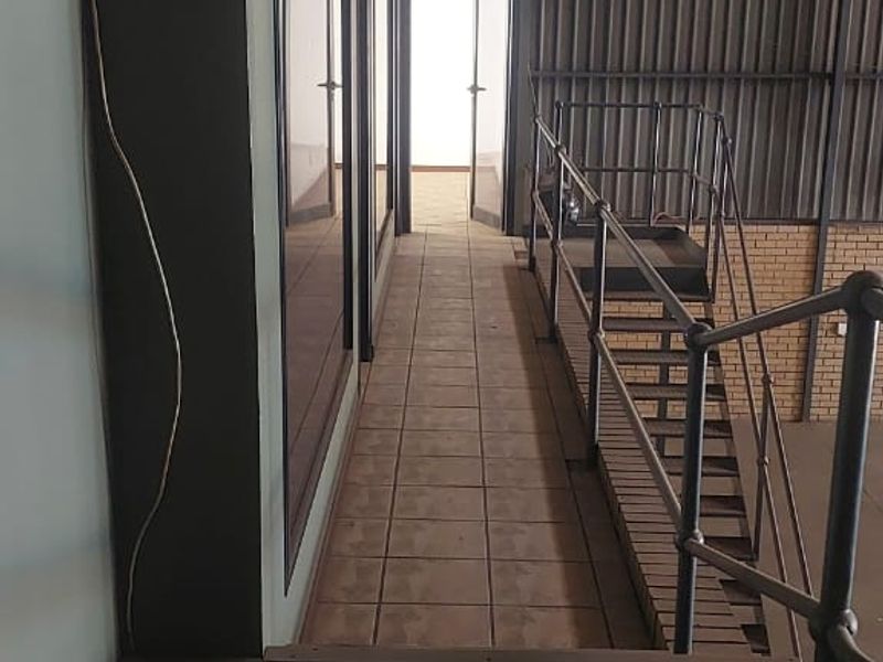 An industrial mezzanine hallway featuring a metal staircase, tiled flooring, and corrugated metal wall cladding, leading to an exterior doorway.