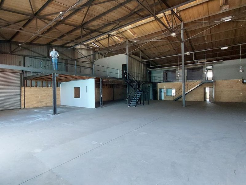 Spacious industrial warehouse featuring a steel mezzanine level, concrete flooring, exposed roof trusses, and a large roller shutter door for easy access.