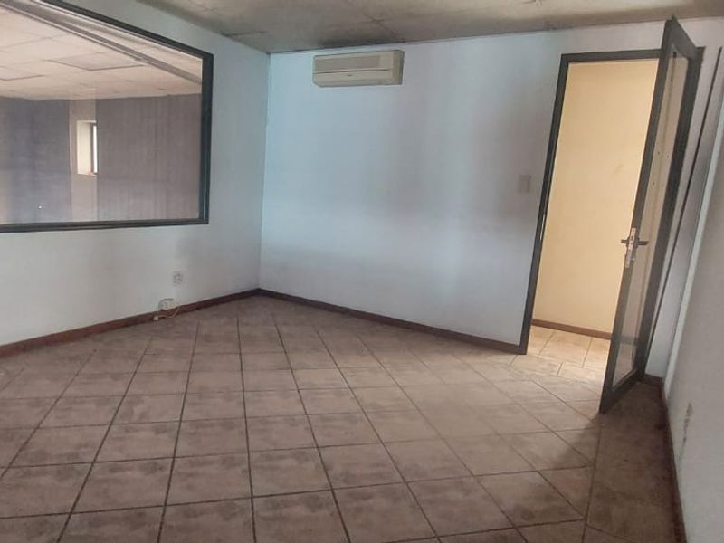 An empty office space featuring tiled flooring, a wall-mounted air conditioning unit, and an internal glass partition window looking into an adjacent area.