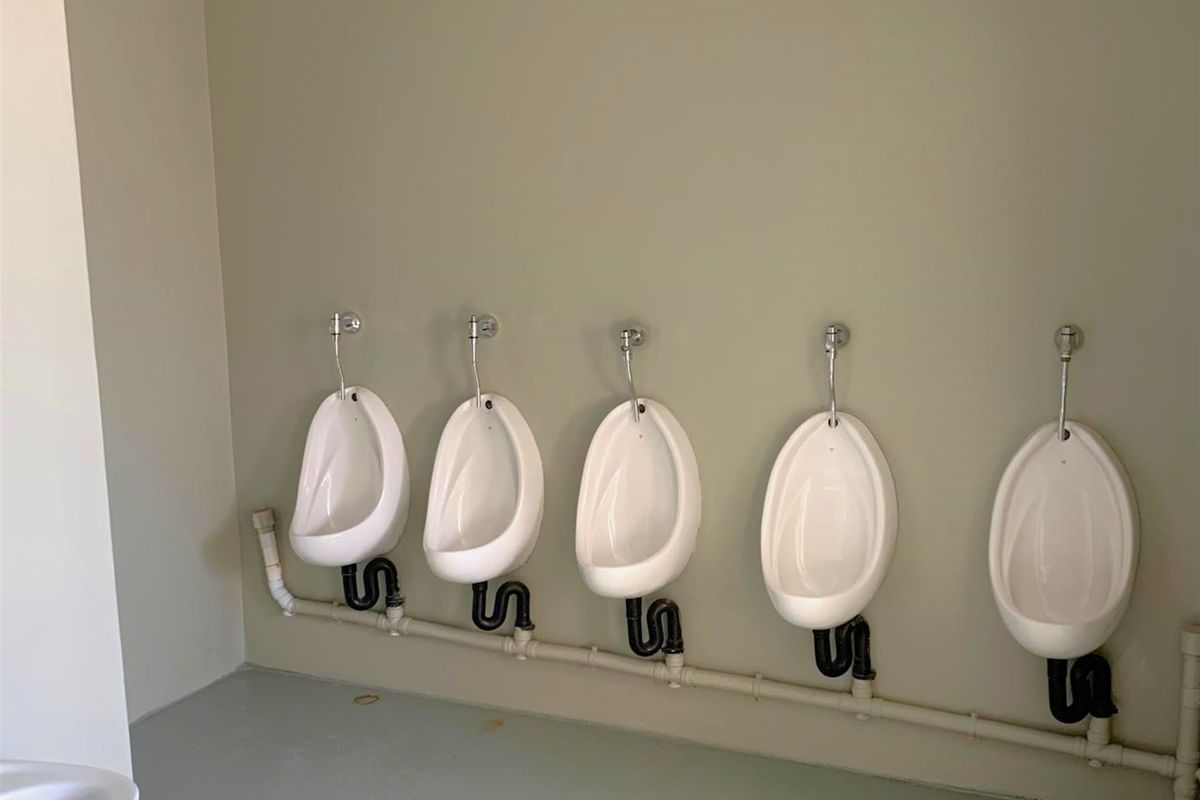 A commercial bathroom facility featuring a row of five wall-mounted white ceramic urinals with exposed plumbing and waste pipe infrastructure.