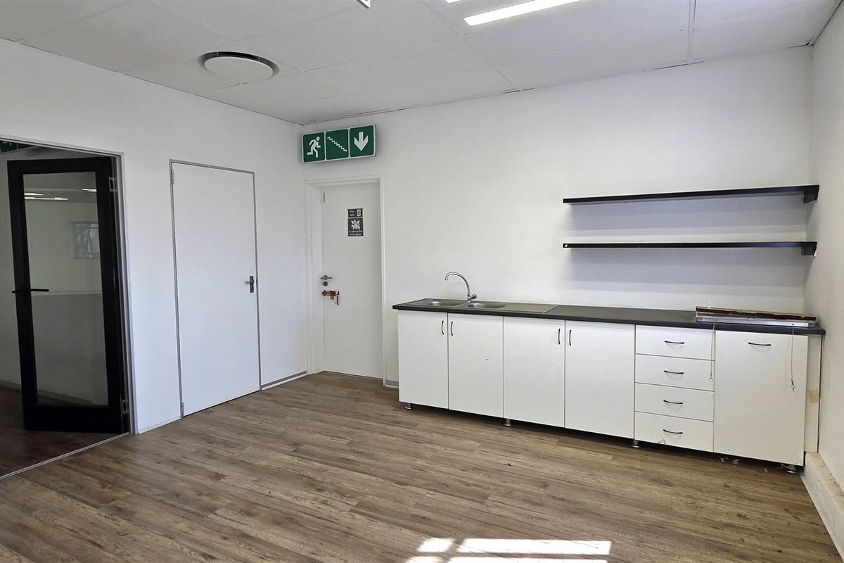 A clean, functional office kitchenette featuring white cabinetry, a stainless steel sink, laminate countertops, and floating shelving in a bright, open workspace.