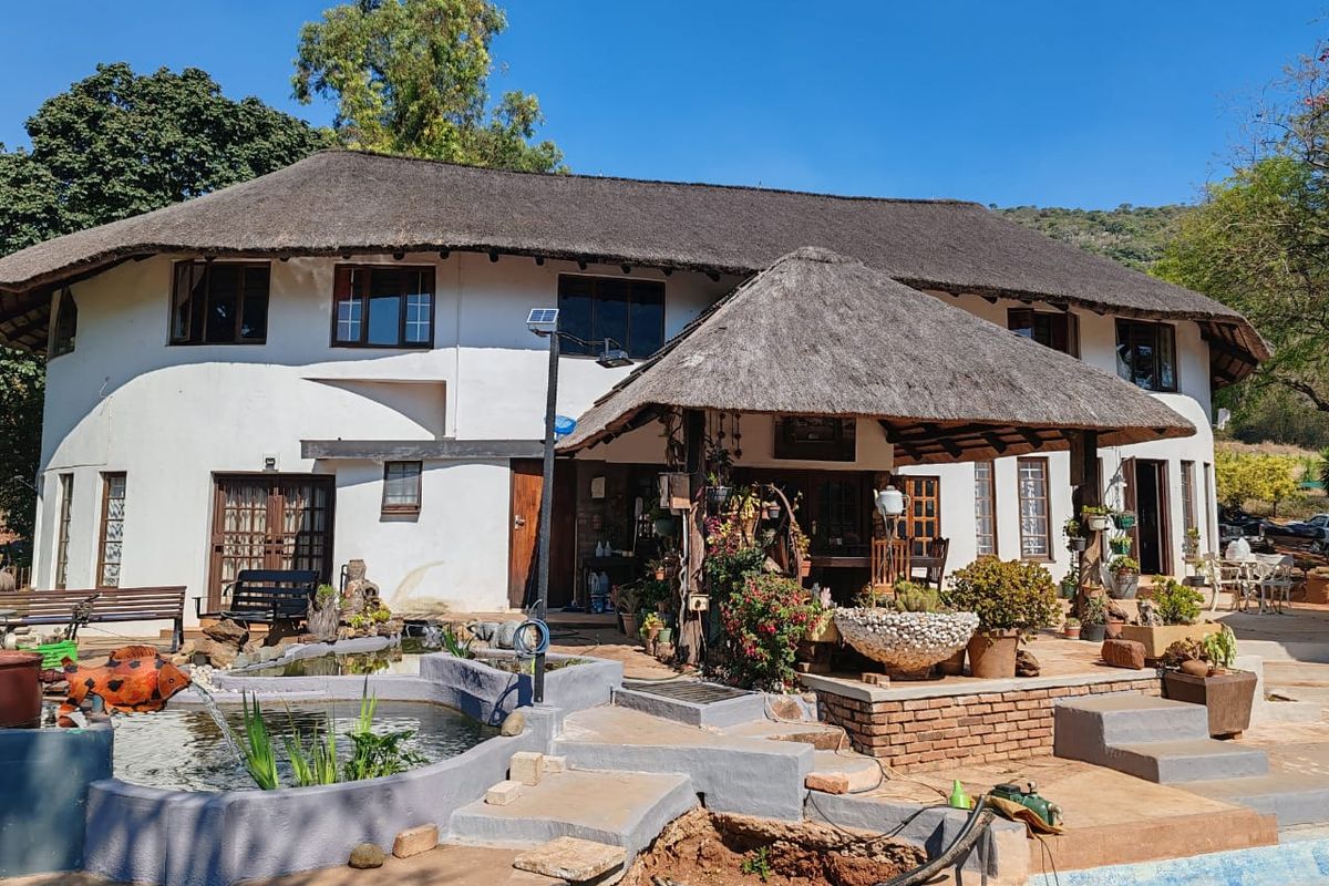 Rear view of a farmhouse featuring a thatched roof, covered patio area, and a garden pond, set against a natural landscape.