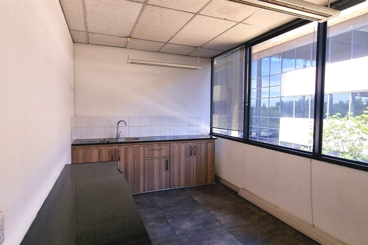 A functional office kitchenette featuring dark stone countertops, wood-finish cabinetry, a stainless steel sink, and large windows providing natural light.