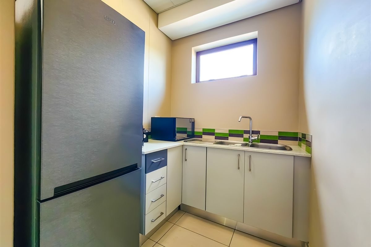 A compact office kitchenette featuring built-in cabinetry, a stainless steel sink, tiled splashback, and space for a refrigerator.