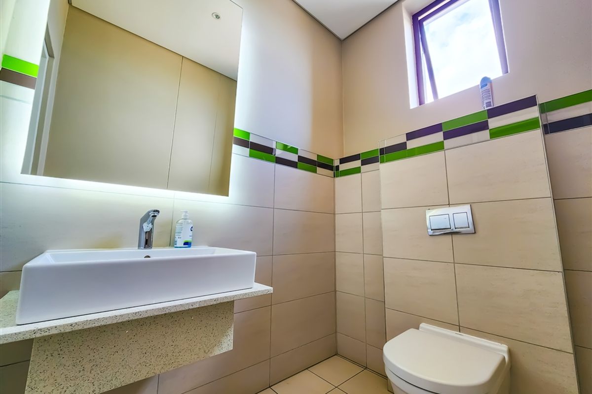 Modern office restroom featuring a wall-mounted basin, integrated mirror with lighting, and a concealed cistern toilet with tiled finishes.