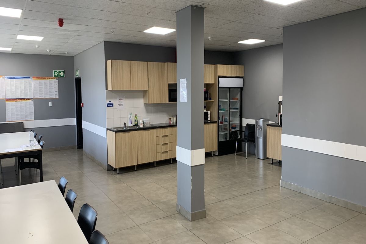 A functional industrial kitchenette area featuring light wood cabinetry, dark countertops, tiled flooring, and recessed ceiling lighting within an open-plan office space.