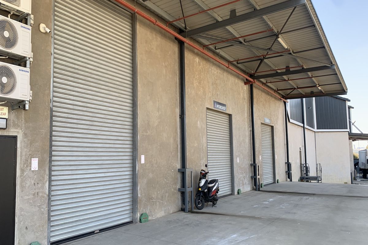 Industrial warehouse exterior featuring multiple roller shutter doors, covered loading bay area, and wall-mounted air conditioning units.