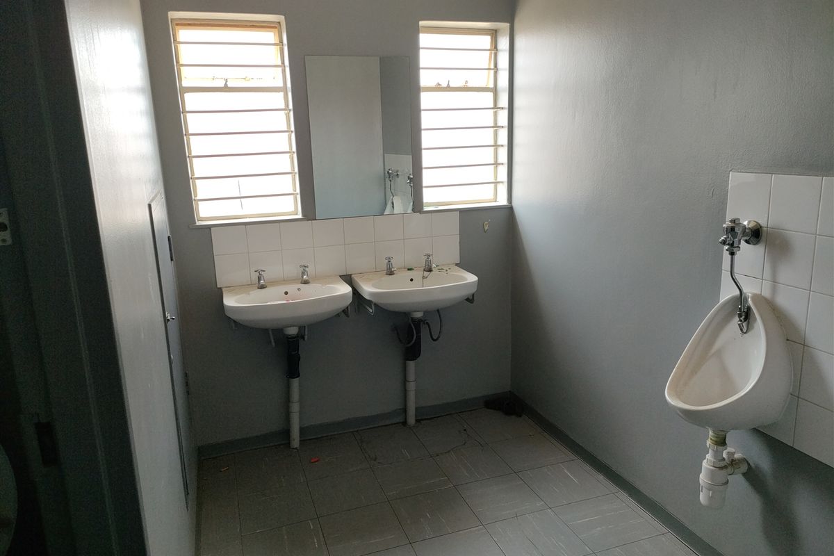 Industrial restroom facility featuring dual wall-mounted ceramic basins, a single wall-mounted urinal, and tiled flooring with louvered windows.