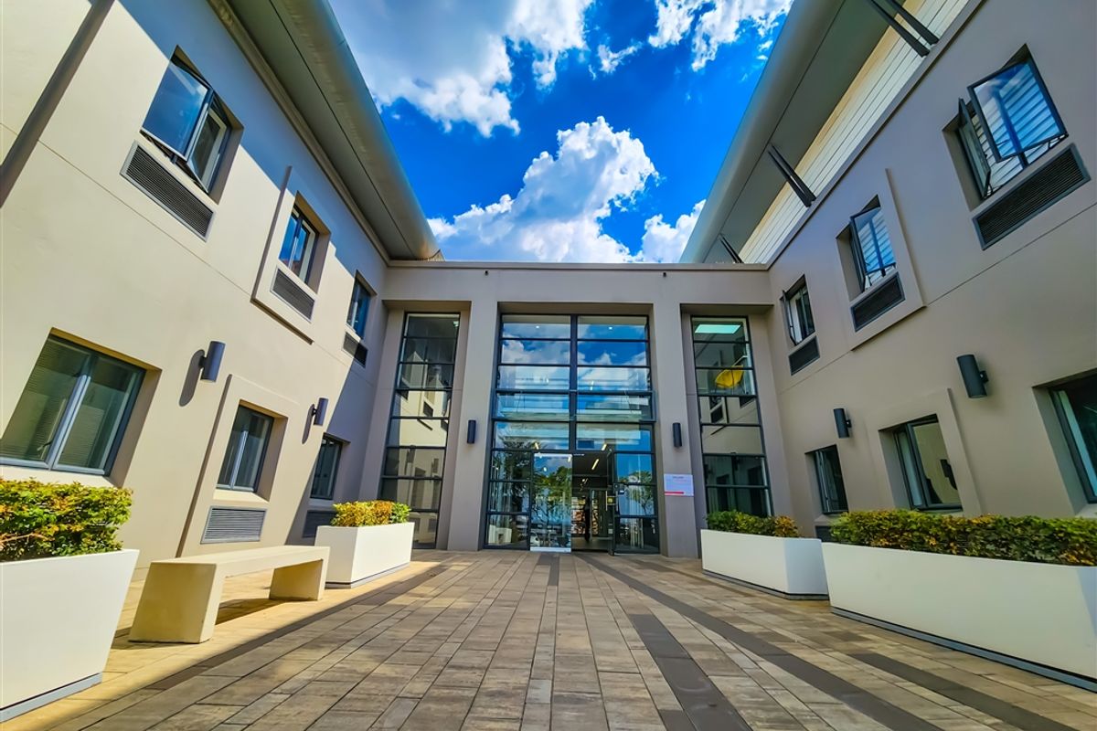 A modern office building courtyard featuring paved walkways, integrated planters, and a large glass-fronted entrance facade.