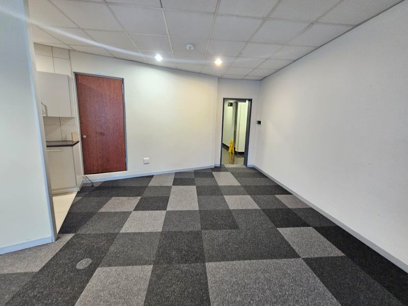 An empty office space featuring a suspended ceiling, recessed lighting, and modern checkered carpet tiles, including a small integrated kitchenette area.