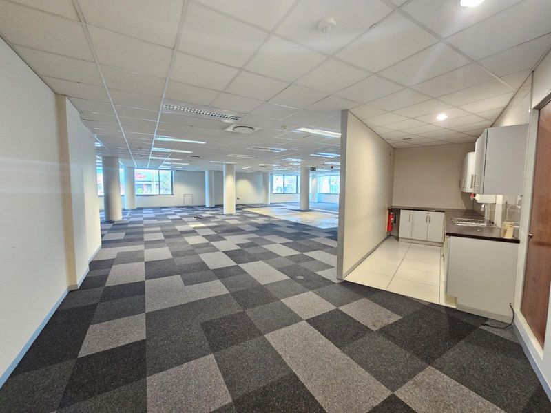 Spacious open-plan office area featuring a suspended ceiling, carpet tiles, structural columns, and a functional kitchenette with built-in cabinetry.