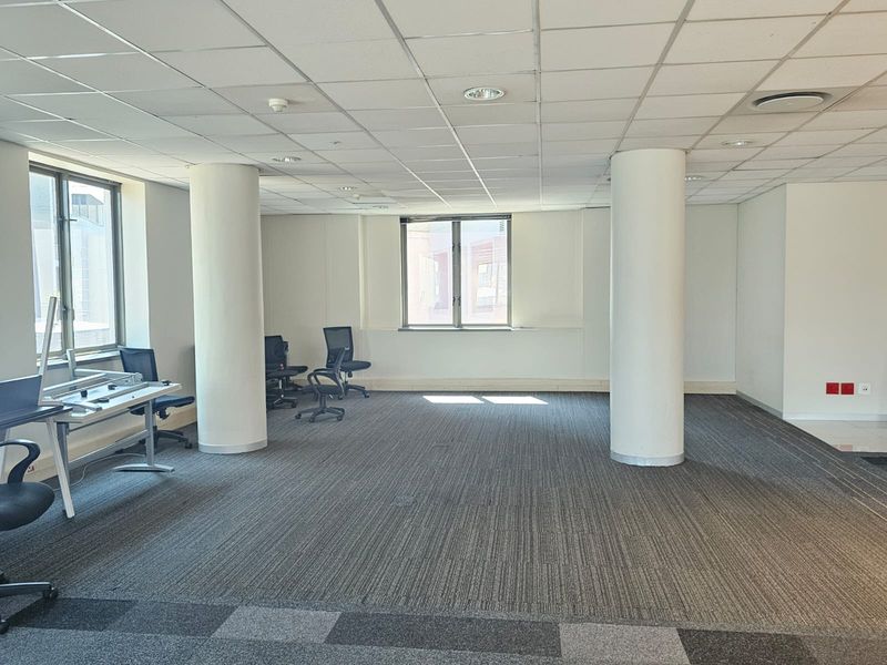 Open-plan office space featuring structural columns, suspended ceilings with recessed lighting, and carpeted flooring, offering a functional workspace environment.