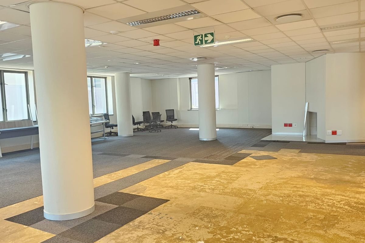 Spacious open-plan office area featuring structural columns, suspended ceilings with recessed lighting, and a mix of carpeted and exposed flooring.