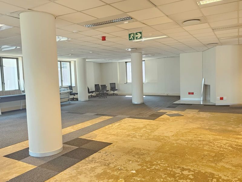 Spacious open-plan office area featuring structural columns, suspended ceilings with recessed lighting, and a mix of carpeted and exposed flooring.