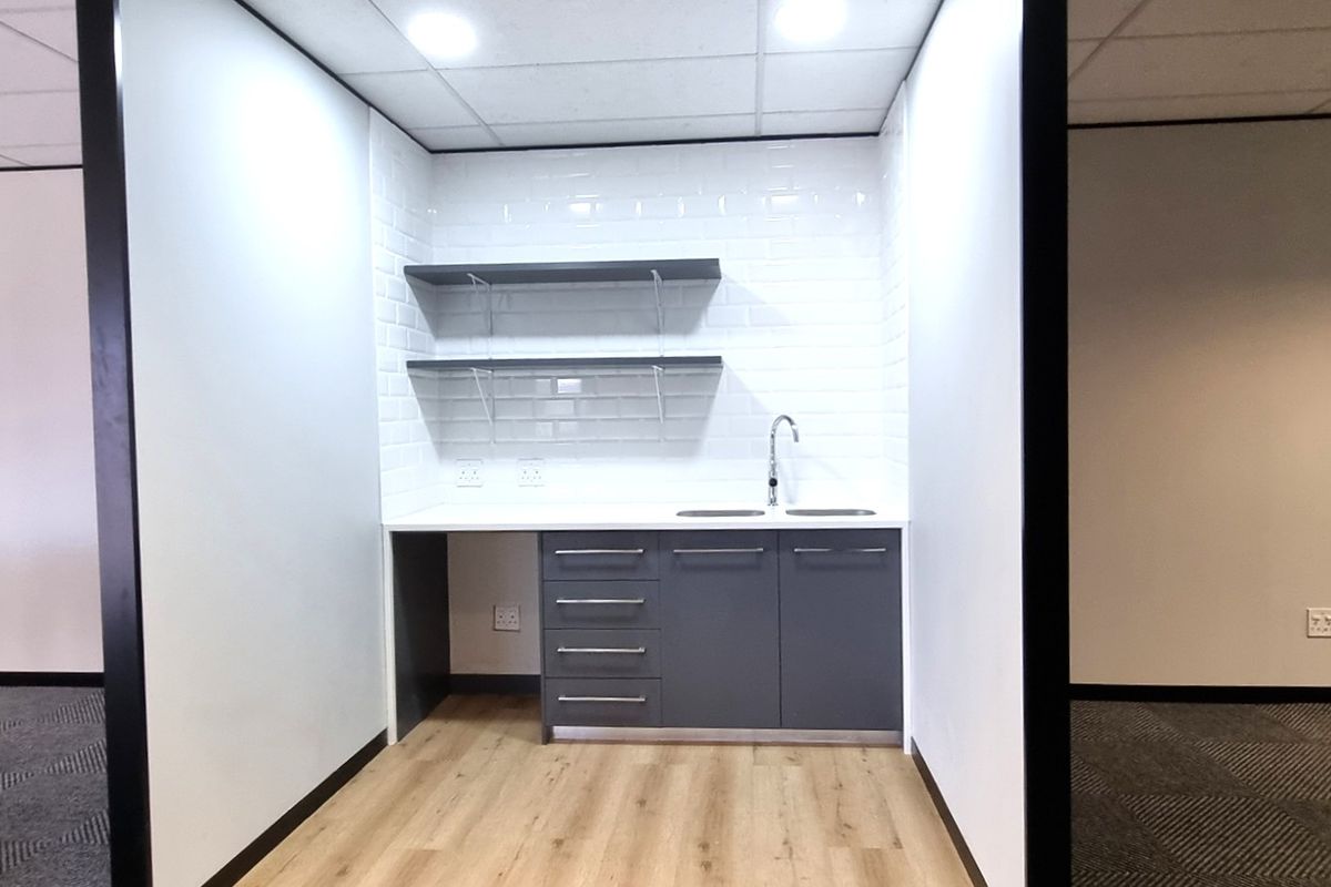 A modern office kitchenette featuring grey cabinetry, white subway tile backsplash, open shelving, and a stainless steel sink.