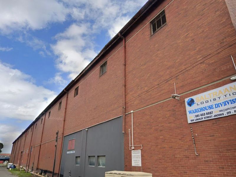 Side elevation of a large industrial warehouse building featuring red brick construction, external utility infrastructure, and commercial signage.