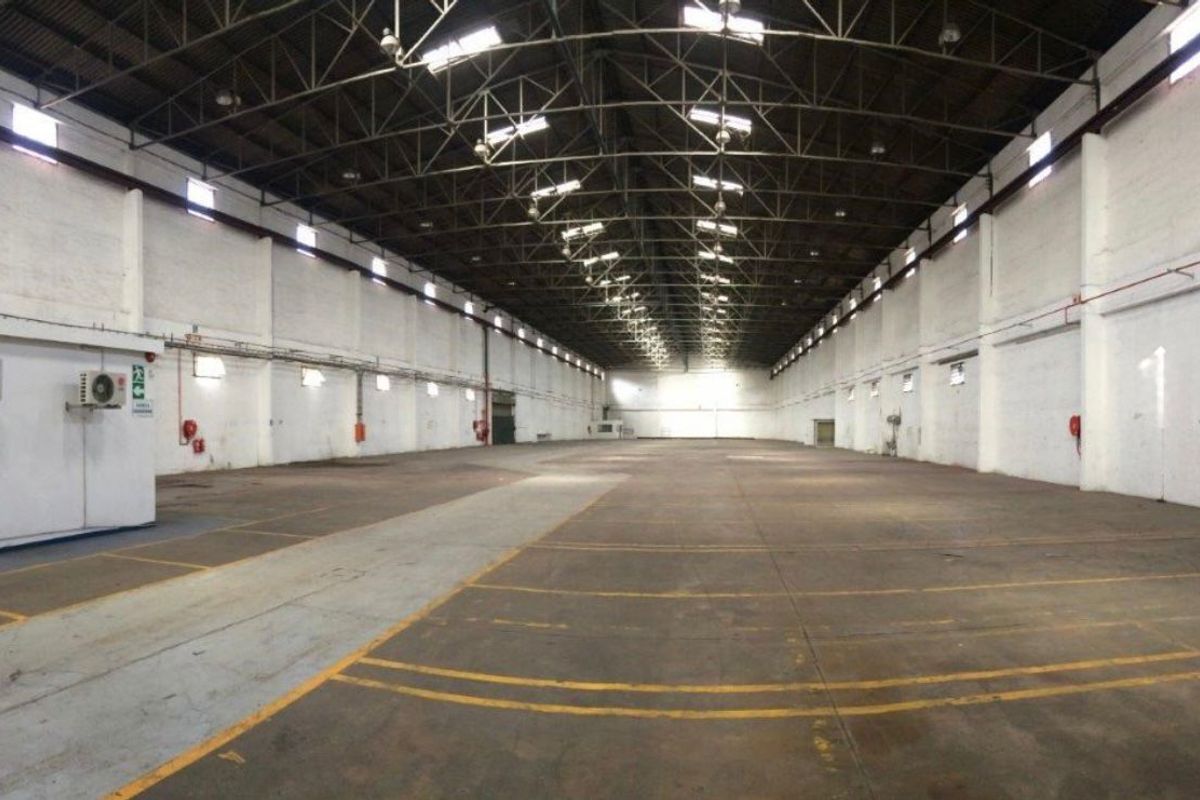 Large open-plan industrial warehouse space featuring high ceilings with exposed steel trusses, concrete flooring, and wall-mounted utility fixtures.