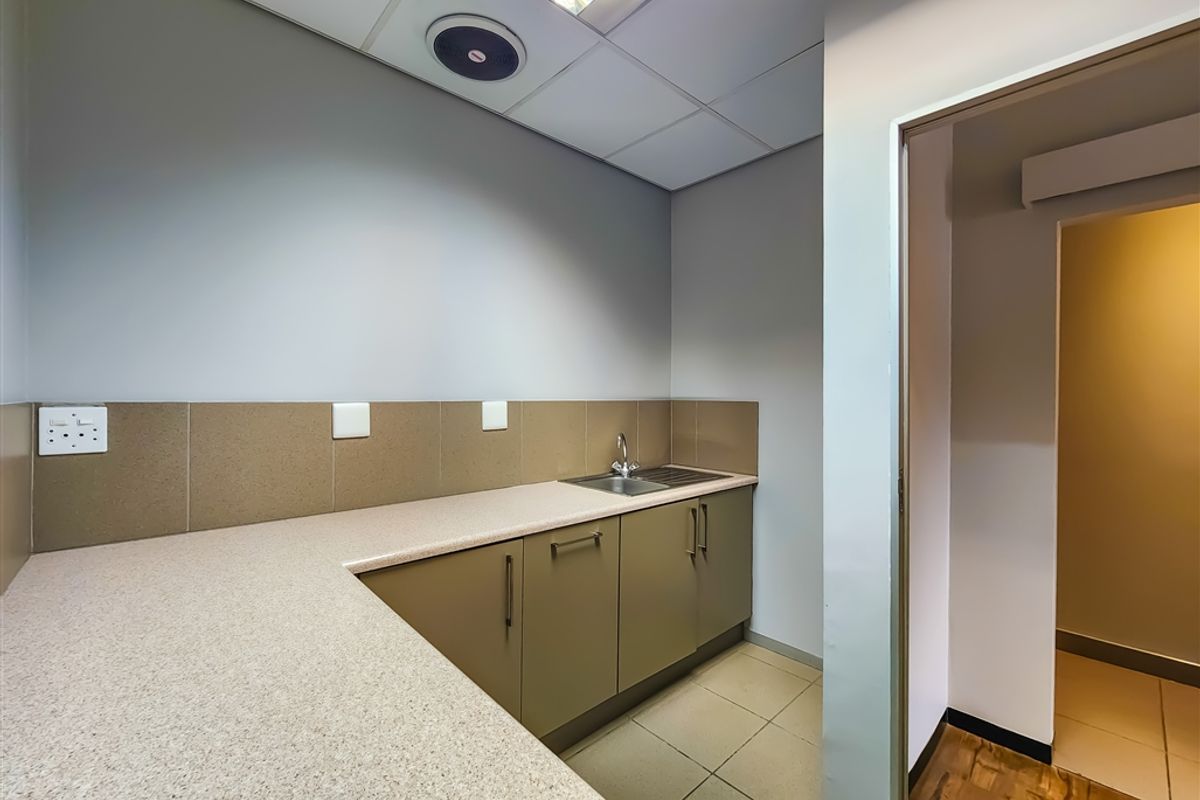 A functional office kitchenette featuring built-in cabinetry, a laminate countertop, a stainless steel sink, and a suspended ceiling with integrated lighting.