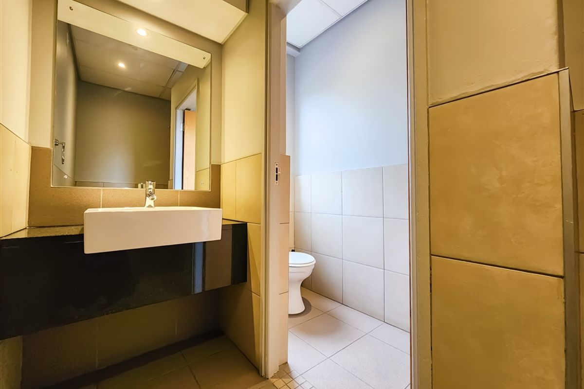 A functional office restroom featuring a wall-mounted basin with a black vanity top, large mirror, and a separate toilet cubicle with tiled finishes.
