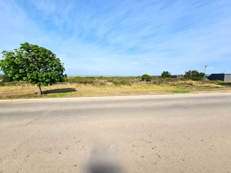A street-level view of a vacant commercial land plot featuring paved road access, perimeter fencing, and open landscape.
