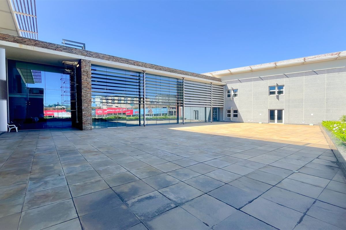 A spacious paved outdoor courtyard area featuring modern glass facade architecture, stone cladding accents, and integrated louvered sun shading systems.