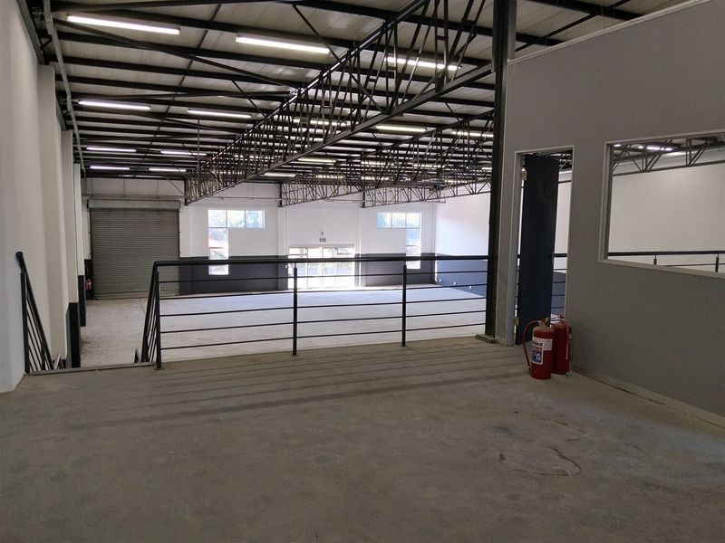 Spacious industrial warehouse interior featuring a mezzanine level, exposed steel roof trusses, roller shutter door, and large windows providing ample natural light.