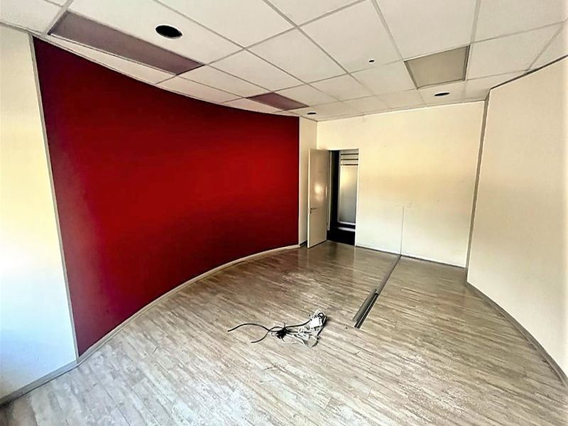 An empty office space featuring a bold red accent wall, suspended ceiling tiles, and light wood-look laminate flooring.