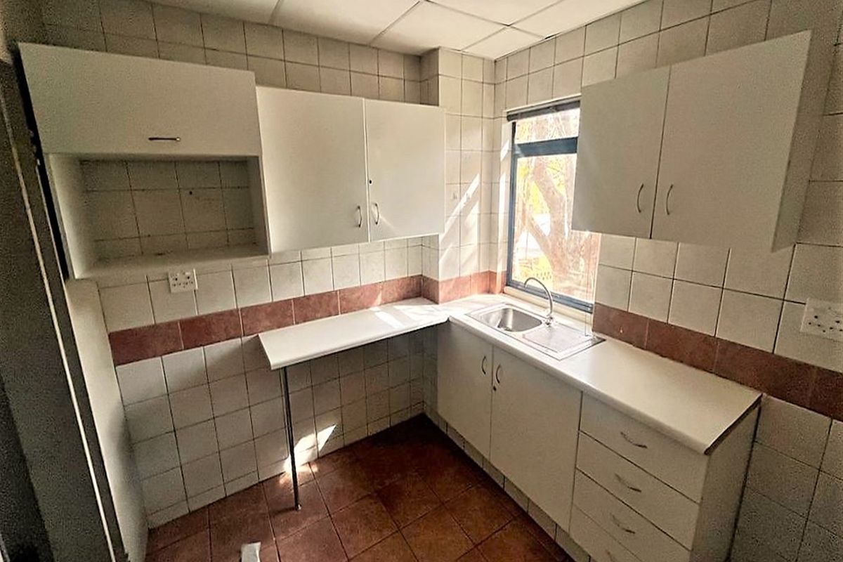 A compact, empty kitchenette featuring built-in white cabinetry, tiled surfaces, and a stainless steel sink, suitable for office staff use.