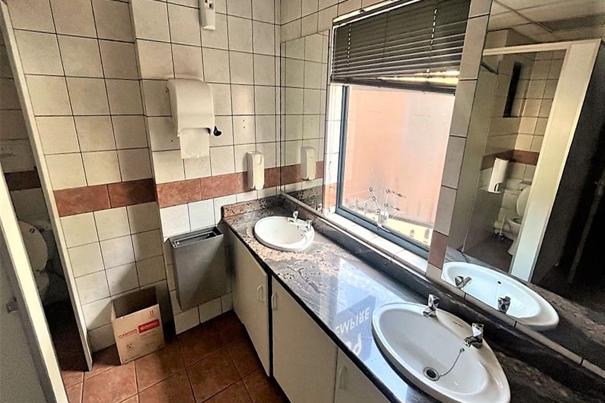 A functional office bathroom featuring dual vanity basins with granite countertops, tiled finishes, and a large mirror, requiring cosmetic updates.