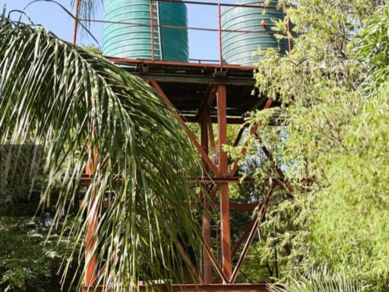 An elevated steel structure supporting two large green water storage tanks, situated within a garden setting.