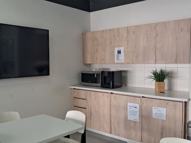 A functional office kitchenette featuring light wood-tone cabinetry, a tiled backsplash, laminate countertops, and a wall-mounted television for presentations or information.