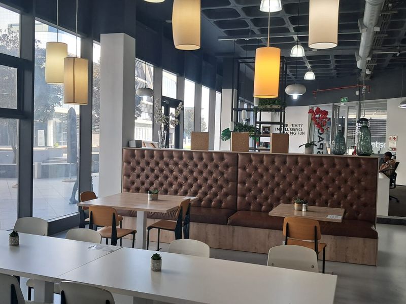 Modern open-plan office breakout area featuring polished concrete floors, floor-to-ceiling windows, exposed architectural ceiling, and integrated tufted leather banquette seating.