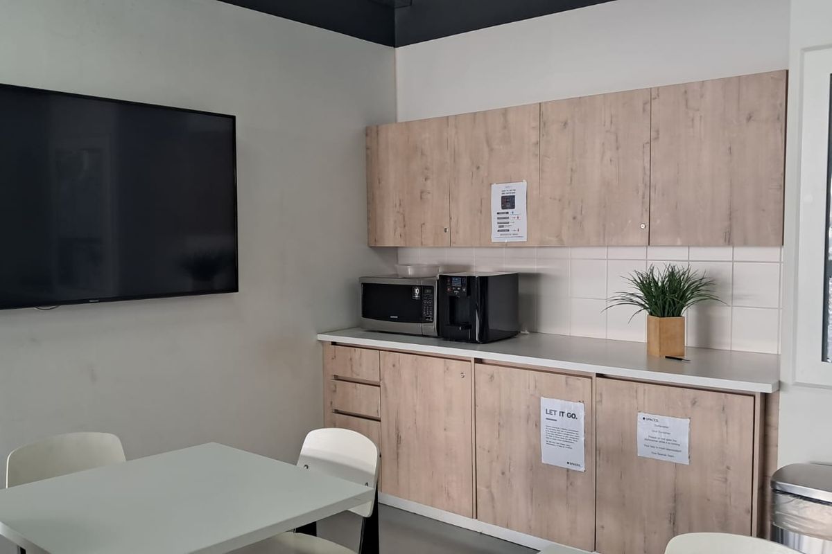 A functional office kitchenette featuring light wood-tone cabinetry, a tiled backsplash, laminate countertops, and a wall-mounted television for presentations or information.